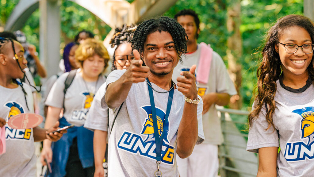 Group of students in UNCG t-shirts. A male in the middle is pointing to the camera with both hands.