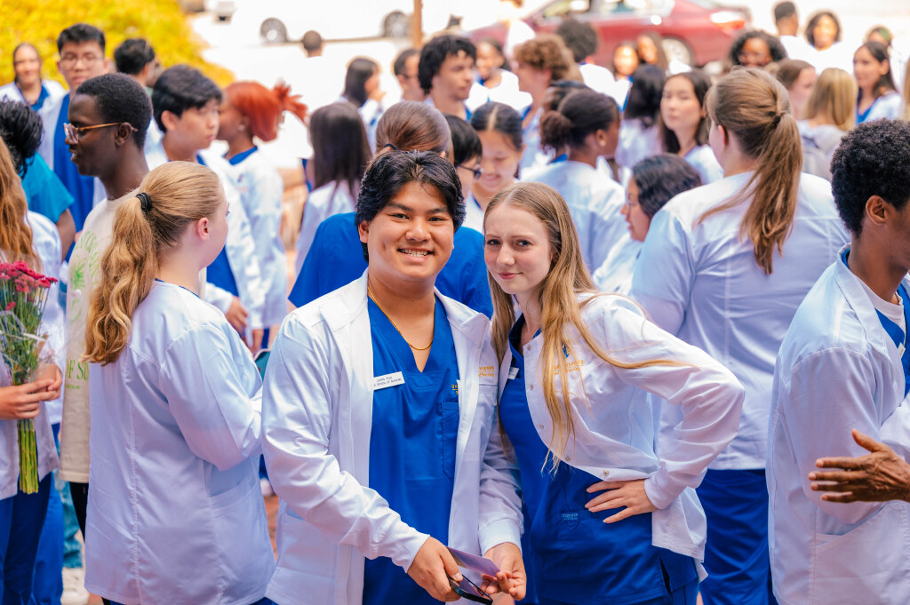 Two nursing students pose for camera in lab coats and scrubs among other nurses who are mingling in background.