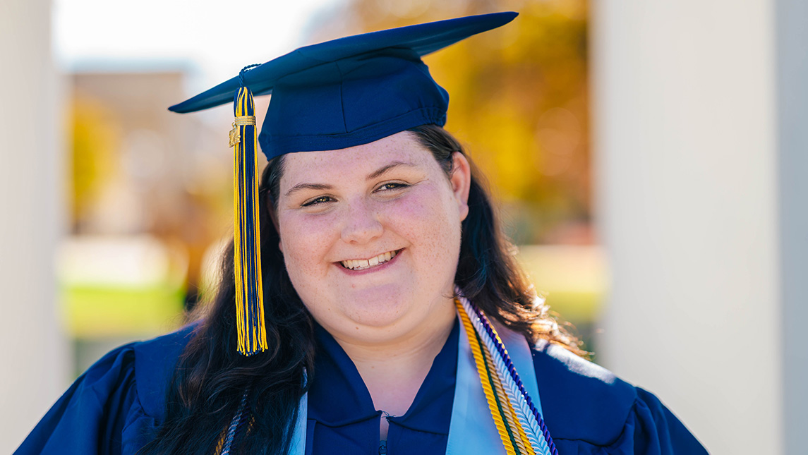 Close-up of a student in cap and gown standing in front of the clock tower at UNCG.