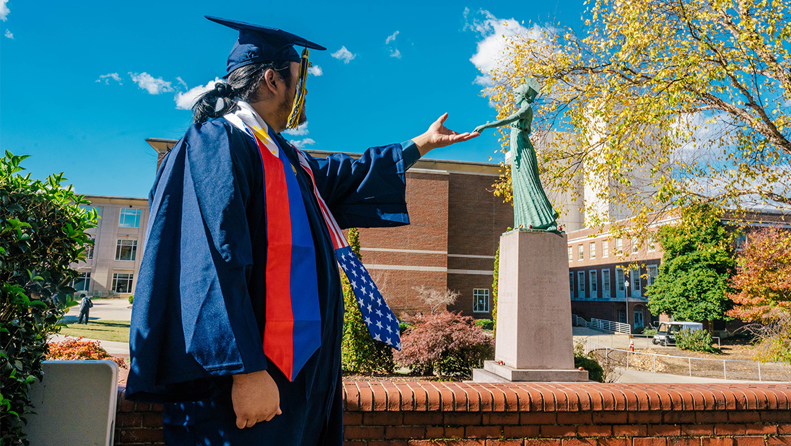 Elijah Resuello holds out his hand toward the UNCG Minerva statue.