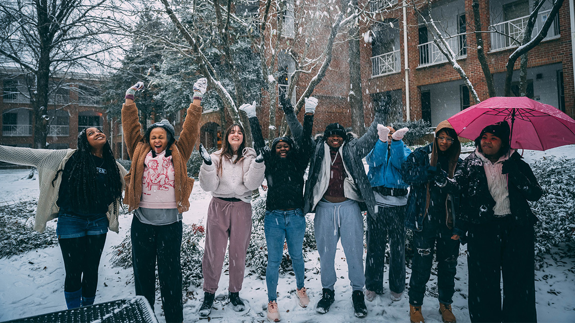 A group of students stand outside of a residence hall and cheer as snow falls.