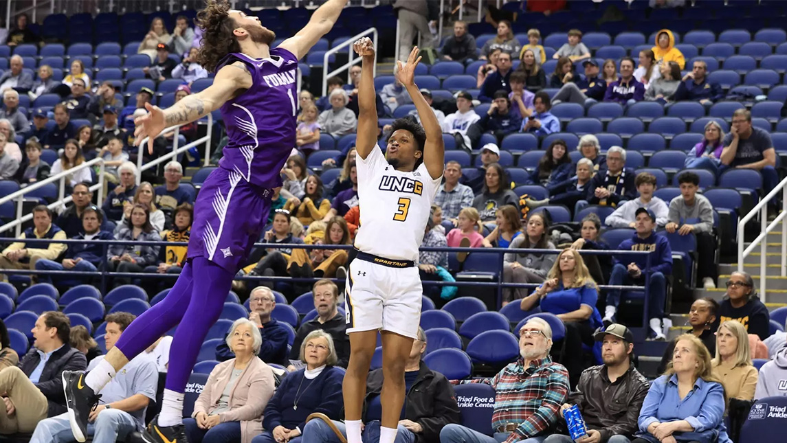 UNCG basketball player Kenyon Giles jumps to block a shot by a Furman player.