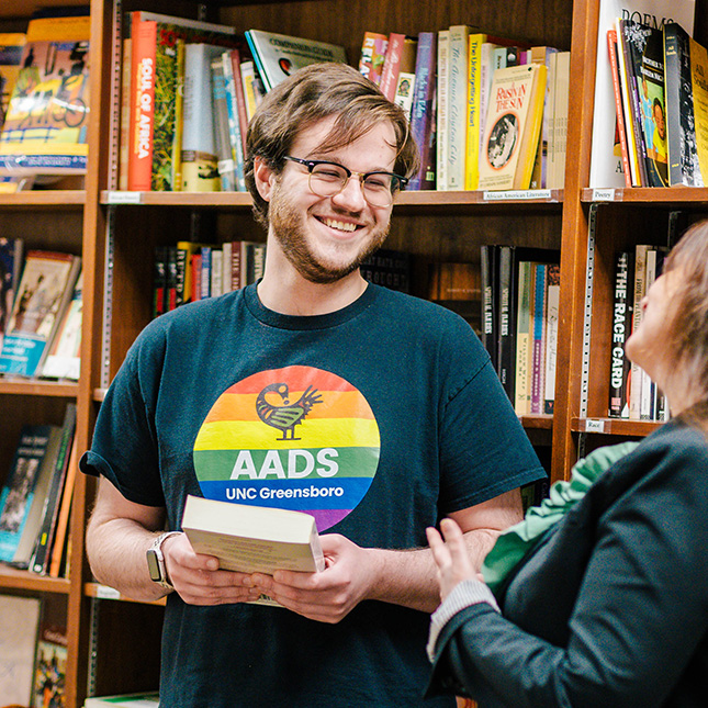 Student in a black t-shirt with a rainbow colored AADS logo talks to a professor in a library with a big smile.