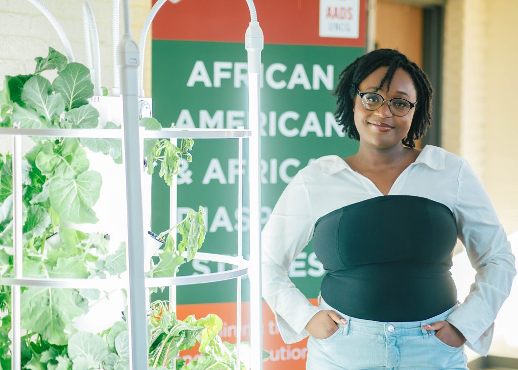 Student stands beside a hydroponic garden with lettuce growing in it and a AADS banner behind her.