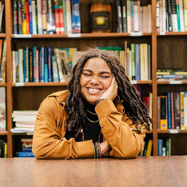 Student sits at a table in a library in a casual pose.