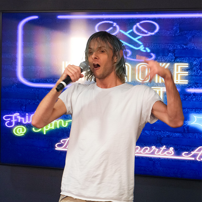 Student sings on a microphone in front of a screen with karaoke graphics in the esports arena.