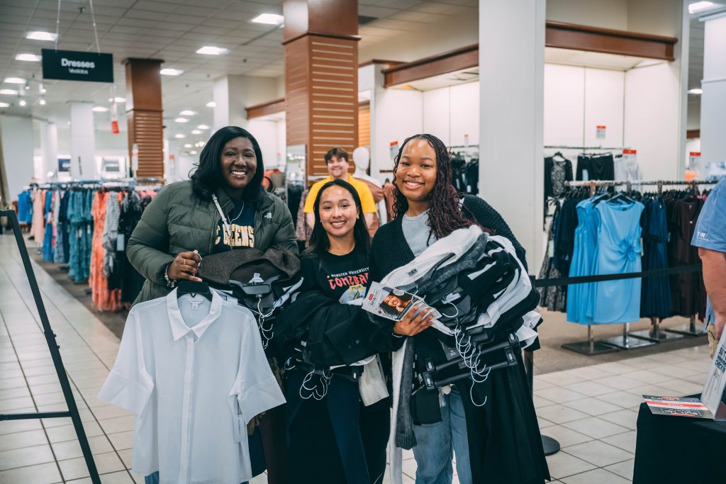 Three students stand with arms loaded down with business clothes.